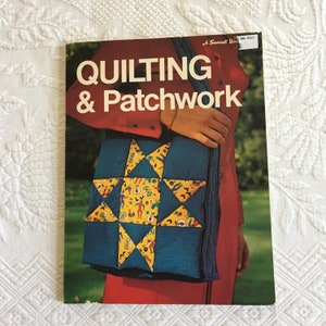 Pode incluir: Um livro intitulado "QUILTING & Patchwork" com uma bolsa de patchwork vermelho, branco e azul na capa. A bolsa é um design de estrela e está sendo segurada por uma pessoa que está vestindo uma camisa vermelha.