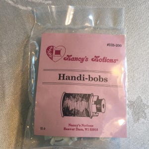 May include: A packet of Nancy's Notions Handi-bobs, sewing accessories. The pink label features a heart with a spool of thread and the text "Nancy's Notions" and "Handi-bobs". The package also includes the text "Beaver Dam, WI 53916".