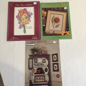 Op de afbeelding: Drie kruissteekpatronen, een met een boeket van lila's, tulpen en narcissen, een ander met een zonnebloem en de laatste met een stilleven met fruit. De patronen zijn gedrukt op papier met een witte achtergrond en zijn getiteld "Lilacs, Tulips, and Daffodils", "Flowers for You" en "Fruit Ambrosia".