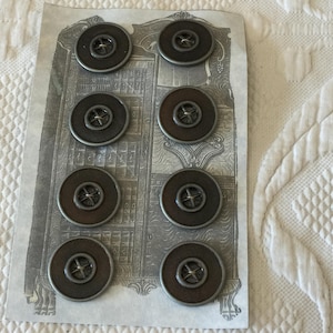 Op de afbeelding: Acht zilveren en bruine metalen knopen met een kruisontwerp in het midden. De knopen zijn gerangschikt in twee rijen van vier op een witte achtergrond.