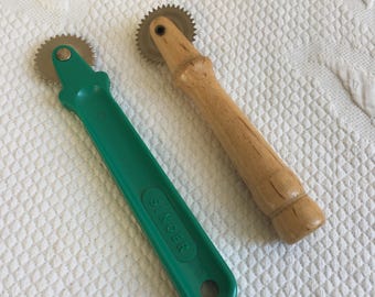 Puede incluir: Dos ruedas de trazado de costura. Una tiene un mango de plástico verde con la palabra "SINGER" impresa. La otra tiene un mango de madera. Ambas tienen una rueda metálica circular dentada.