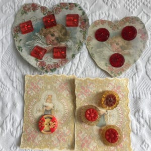 May include: A collection of vintage Valentine's Day ephemera, including heart-shaped cards with cherub illustrations and red glass buttons. The cards feature floral and lace designs. Some cards have the text "A Token of Love".