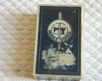 Puede incluir: Una caja rectangular blanca con una etiqueta azul. La etiqueta tiene un logotipo en blanco y negro de una máquina de coser con el texto "Needle Works" alrededor.
