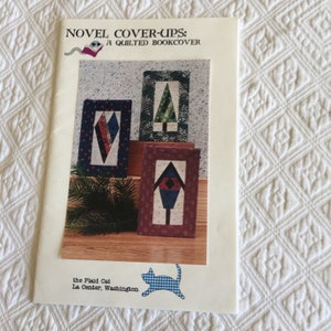 May include: A pamphlet titled "Novel Cover-Ups: A Quilted Bookcover" with a picture of three quilted book covers. The book covers are decorated with a diamond pattern, a Christmas tree, and a geometric design. The pamphlet is from "the Plaid Cat" in La Center, Washington.