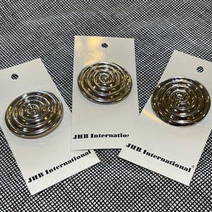 Könnte beinhalten: Drei silberfarbene, spiralförmige Zierpins auf weißen Karten. Auf jeder Karte steht der Text "JHB International". Der Hintergrund ist ein schwarz-weiß gemusterter Stoff.