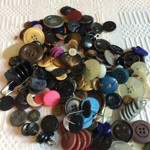 May include: A collection of various buttons in different sizes, shapes, and colors. The assortment includes buttons in shades of black, brown, white, pink, and blue. Some buttons have safety pins attached.