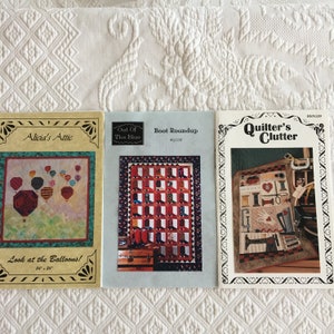 Könnte beinhalten: Drei Quilting-Musterbücher mit bunten Umschlägen. Das erste Buch trägt den Titel "Alicia's Attic" und zeigt ein farbenfrohes Heißluftballon-Design. Das zweite Buch trägt den Titel "Out Of The Blue" und zeigt ein rot-weißes Quilt-Design. Das dritte Buch trägt den Titel "Quilter's Clutter" und zeigt ein farbenfrohes Quilt-Design mit einem Näh-Thema.
