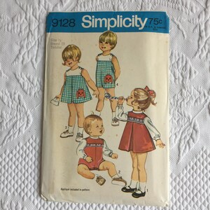 Può includere: Una busta per cartamodello Simplicity 9128 per bambini piccoli, taglia 1/2, torace 19. La copertina presenta illustrazioni di bambini che indossano vari abiti, tra cui un vestito a quadretti verdi, una tutina e un vestito rosso a maniche lunghe.