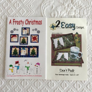 May include: Two quilting pattern books with a Christmas theme. The first book is titled "A Frosty Christmas" and features a snowman and Christmas trees. The second book is titled "2 Easy Designs" and features a set of four beverage mats with a Christmas tree design. Each mat is 6 inches by 10 inches.