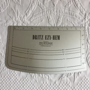 May include: A silver-colored metal Dritz Ezy-Hem sewing gauge featuring black printed text and measurement markings. The tool displays the text Dritz Ezy-Hem, Designed by Edna Bryte Bishop, Originator of the Bishop Method of Clothing Construction.