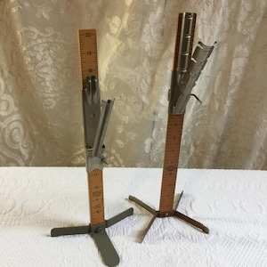 May include: Two vintage skirt markers with wooden vertical rulers featuring metric measurements up to 50 cm. Each tool has a metal base, a sliding metal guide for marking hemlines, and a built-in pin cushion mechanism at the top.