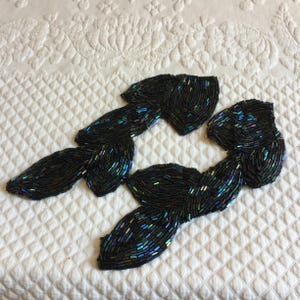 Pode incluir: Um par de enfeites de contas pretas com acentos azuis e verdes iridescentes. Os enfeites têm a forma de folhas e estão conectados entre si.