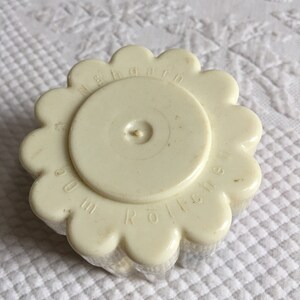 May include: White plastic yarn roller with a flower-shaped design. The roller has the text "N&auml;hgarn Rolle" printed on it.