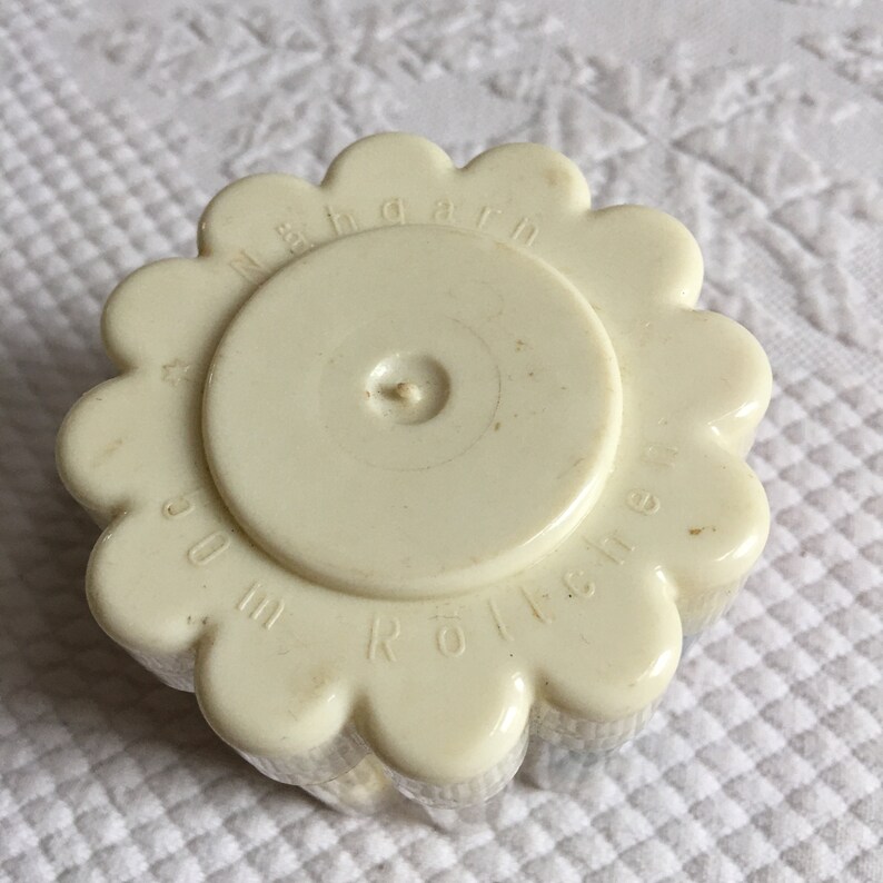 May include: White plastic yarn roller with a flower-shaped design. The roller has the text "N&auml;hgarn Rolle" printed on it.