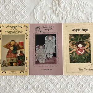 May include: Three craft patterns for making angels. The first pattern is called "Fruitfull Hands - Sweet as an Angel" and features a red and white plaid angel with a green shawl. The second pattern is called "Allison's Angels" and features a country folk angel with a blue and white dress. The third pattern is called "Angela Angel" and features a small angel with a red and white dress and a brown hair bow.