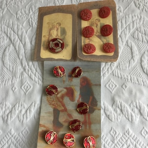 Puede incluir: Un surtido de botones vintage en varios diseños y colores. La imagen presenta botones rojos con patrones en relieve, botones rojos y dorados con detalles de alambre y un botón con un diseño geométrico. Los botones se muestran sobre papel vintage con ilustraciones.