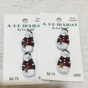 May include: Two cards each displaying two hand-painted snowman pins. The snowmen wear burgundy coats, green scarves, and red-and-green hats. Each snowman holds a gingerbread man. The cards read "A 3-D HOLIDAY by La Mode."