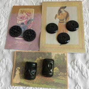 May include: An assortment of vintage black buttons and carved beads are displayed on antique postcards. The buttons are round with intricate designs, while the beads are rectangular with carved faces. The postcards feature illustrations and the words "Easter Greetings".