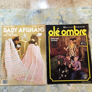 以下が含まれることがあります： 2冊のヴィンテージ手芸本。左の本は「Baby Afghans to Knit and Crochet」というタイトルで、ピンクと白のシェブロン柄のブランケットとテディベアが写っています。右の本は「Olé Ombre Knits and Crochets」というタイトルで、3人の人物が写っています。
