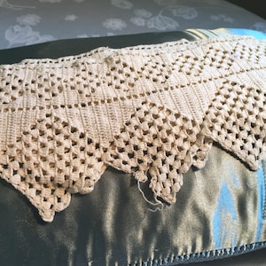 May include: Cream-colored, open-weave crochet doily with a scalloped edge. The handmade textile features a geometric pattern and is displayed on a satin pillow. The doily's texture and design suggest a vintage or antique aesthetic.