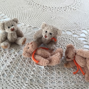 Op de afbeelding: Vier kleine, pluche teddyberen in verschillende poses. De beren zijn voornamelijk bruin en bruin, met zwarte ogen en oranje touwaccenten. Ze zijn gerangschikt op een crèmekleurig, gehaakt oppervlak.