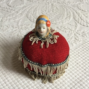 Vintage Pin Cushion. Porcelain Doll Head with Red Woven Wool Look Fabric Ball with Braided Fringe Around Neck and Twice Around String Ball.