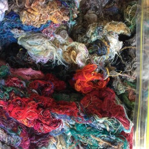 May include: A close-up of a pile of colourful yarn scraps, including shades of red, orange, green, blue, and white. The yarn is tangled and messy, creating a textured and abstract image.