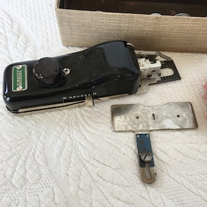 Puede incluir: Una máquina de coser negra y plateada con una etiqueta verde que dice "Greist" y un dial plateado con los números del 1 al 6. La máquina está sobre una superficie blanca con una placa de metal plateada y una pequeña herramienta plateada con un mango azul.
