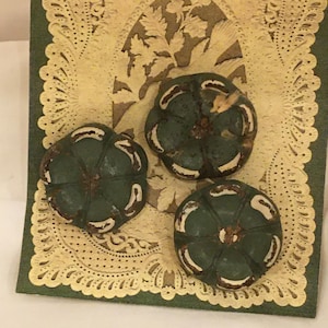 May include: Three dark green, pumpkin-shaped decorative objects with white accents and a distressed finish. They rest on a cream-colored, intricately cut paper with a green border, suggesting a fall or harvest theme.