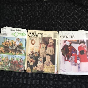 Op de afbeelding: Drie vintage knutselpatroonpakketten. De pakketten bevatten illustraties van handgemaakte poppen en knuffeldieren. De merken zijn Simplicity en McCall's Crafts. De patronen lijken voor naaiprojecten te zijn.