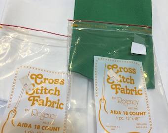 Op de afbeelding: Twee pakketten witte Aida-cross stitch stof, een groen en een wit. Elk pakket bevat een stuk stof dat 30,5 cm bij 45,7 cm is. De pakketten zijn gelabeld "Cross Stitch Fabric by Regency Mills Inc. Aida 18 Count 1 pc. 12"x18""
