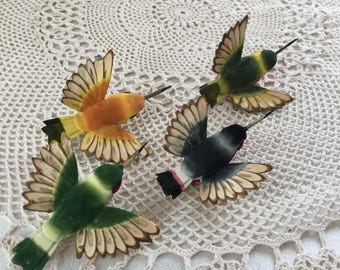 May include: Four colorful hummingbird ornaments with outstretched wings. The birds feature shades of orange, green, black, and yellow with detailed wing patterns. Each has a long, slender beak. The ornaments are arranged on a cream-colored, crocheted surface.