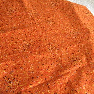 May include: Orange fabric featuring a repeating pattern of thin, squiggly red lines and scattered dashes in yellow, black, and white. The fabric has a slightly wrinkled texture, suggesting it is a soft material.