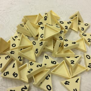 May include: A collection of off-white, triangular plastic game pieces scattered together. Each piece features black printed numbers, including digits zero through five, on its flat surface. The pieces have raised edges.