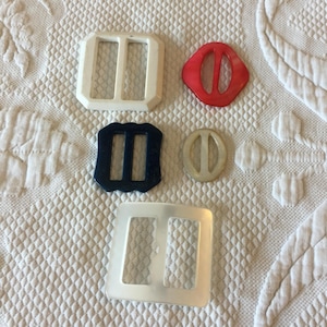 Puede incluir: Cinco hebillas de cinturón de plástico en varios colores y formas. Las hebillas son blancas, rojas, azules y transparentes. Todas tienen forma de rectángulo con un agujero en el medio.