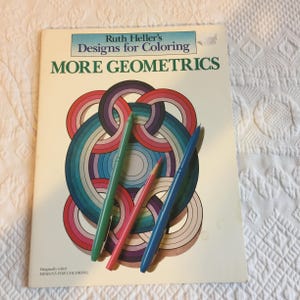 May include: A coloring book titled "Ruth Heller's Designs for Coloring MORE GEOMETRICS" with a colorful geometric design on the cover. Three pens and a pencil in green, pink, and blue rest on the book.