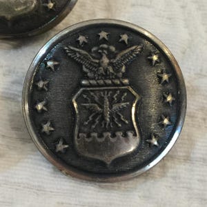 Puede incluir: Botón de metal plateado con el emblema de la Fuerza Aérea de los Estados Unidos. El botón redondo presenta un águila sobre un escudo, rodeada de estrellas. Tiene un aspecto ligeramente envejecido, con un diámetro de unos 2,5 cm.