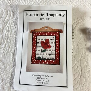 May include: A quilt pattern for a 18 by 21 inch quilt titled "Romantic Rhapsody". The quilt features a red flower with a black and white piano keyboard background. The pattern is from Dinah's Quilts & Accents, 116 Walnut St., Cowan, TN 37318, 931-962-1456.