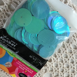 May include: A clear plastic bag filled with iridescent, flat, round, light blue sequins. The bag is partially open, revealing the contents. The sequins are labeled as washable and garment quality. The label also includes the text "sequins" and "flat round 20 mm".