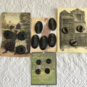 Könnte beinhalten: Eine Sammlung schwarzer Vintage-Knöpfe in verschiedenen Formen und Größen, präsentiert auf antiken Karten. Einige Knöpfe sind oval, andere rund mit aufwendigen Designs. Eine Karte trägt die Aufschrift "Colonial Drapery Fabrics."