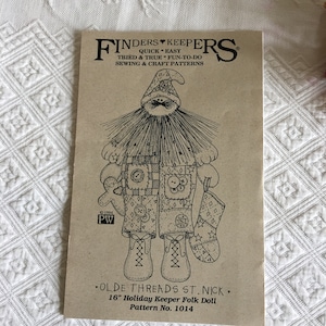 以下が含まれることがあります： 40.6cm のホリデーキーパーフォークドールのパターンの白黒線画。人形はパッチワークの衣装を着ており、片手にストッキングキャップとストッキングを持っています。パターンは「Olde Threads St. Nick」というタイトルで、迅速かつ簡単な縫製とクラフトパターンを作成する会社、Finders Keepers から提供されています。