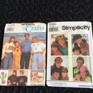 May include: Two vintage Simplicity Crafts sewing pattern envelopes. One features a family in casual attire, the other showcases various hat designs. The envelopes are white with colorful images and text, including the Simplicity logo and pattern numbers.