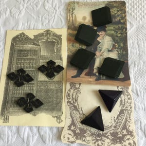 Op de afbeelding: Een assortiment vintage zwarte knopen in verschillende vormen, waaronder vierkanten en ruitvormige ontwerpen. De knopen worden getoond op antieke ansichtkaarten en illustraties, wat een klassieke esthetiek laat zien. De afbeelding is geplaatst tegen een witte, getextureerde achtergrond.