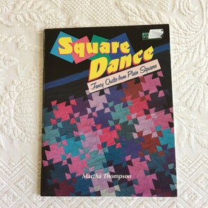 May include: Book cover with the title "Square Dance" in yellow letters on a black background. The subtitle is "Fancy Quilts from Plain Squares". The cover features a colorful quilt pattern with pink, blue, and purple squares.