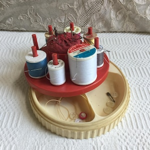 Puede incluir: Un kit de costura vintage con una parte superior roja en capas que contiene carretes de hilo y un alfiletero. La sección inferior es una bandeja de color crema con botones y un imperdible. Los carretes tienen hilo blanco, azul y gris.