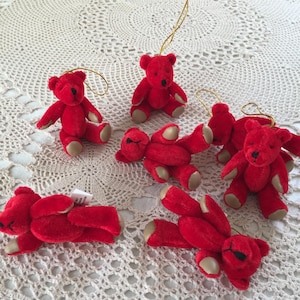 Op de afbeelding: Een verzameling kleine, rode teddybeerornamenten met beige poten en zwarte ogen. Elke beer heeft een gouden draad om op te hangen. De beren staan in verschillende poses, sommige zitten en andere liggen.