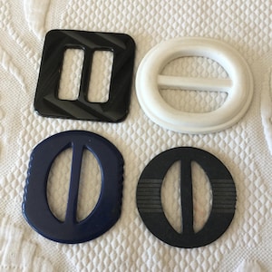 Puede incluir: Cuatro hebillas de cinturón vintage de varias formas y colores. Incluye una hebilla cuadrada negra con líneas diagonales, una hebilla ovalada blanca, una hebilla ovalada azul marino y una hebilla ovalada negra con líneas horizontales.