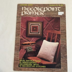 May include: A vintage needlepoint primer booklet with a brown woodgrain cover. The cover features a picture of a white pillow with needlepoint embroidery, a small basket with needlepoint embroidery, and a framed needlepoint picture on a brick wall. The text on the cover reads "Leisure Arts Needlepoint Primer Leaflet 74 $1.50" and "Basic Instructions in Needlepoint Techniques with 12 Decorative Stitches".