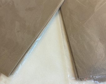 May include: Two pieces of tan cross-stitch fabric in clear plastic packaging, with a white cross-stitch fabric in the background. The tan fabric has a visible grid pattern, ideal for needlework projects. The plastic packaging is sealed with a pink zipper.