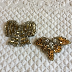 Puede incluir: Dos adornos de tela de color dorado. Uno es un lazo hecho de cuentas y el otro es una forma de mariposa hecha de lentejuelas y cuentas.